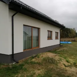 Świeżo otynkowany dom jednorodzinny z ciemną dachówką i brązowymi oknami, widok z boku na trawnik z fragmentami piasku.