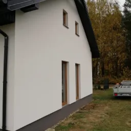 Boczny widok domu jednorodzinnego z białą elewacją i grafitowym dachem, widoczny fragment rynny, dwa małe okna na poddaszu i dwa większe okna na parterze, w tle drzewa z jesiennymi liśćmi...