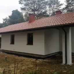 Parterowy dom jednorodzinny z czerwoną dachówką, świeżo po malowaniu elewacji na biało, widoczny z boku na tle drzew.