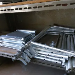 Stos ocynkowanych, metalowych elementów konstrukcyjnych z otworami i mocowaniami, ułożonych na półce w magazynie.