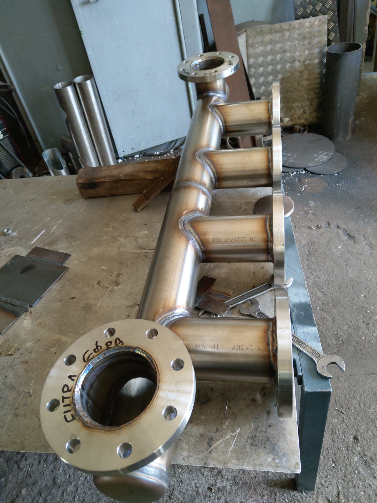 Poskładana konstrukcja rurowa ze stali nierdzewnej z widocznymi spawami i kołnierzami, leżąca na warsztatowym stole, obok narzędzi.