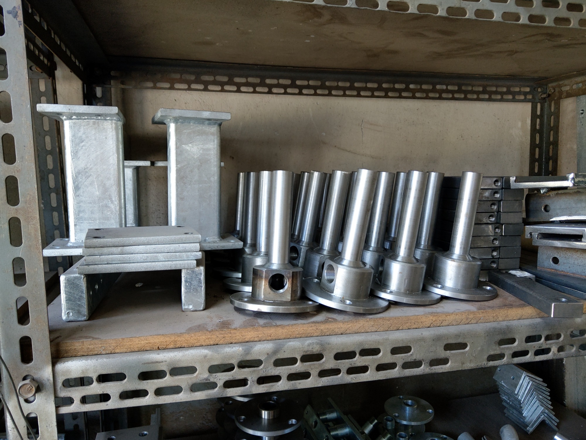 Półki regału warsztatowego wypełnione metalowymi elementami konstrukcyjnymi, w tym kolumny, podstawy z otworami i kątowniki