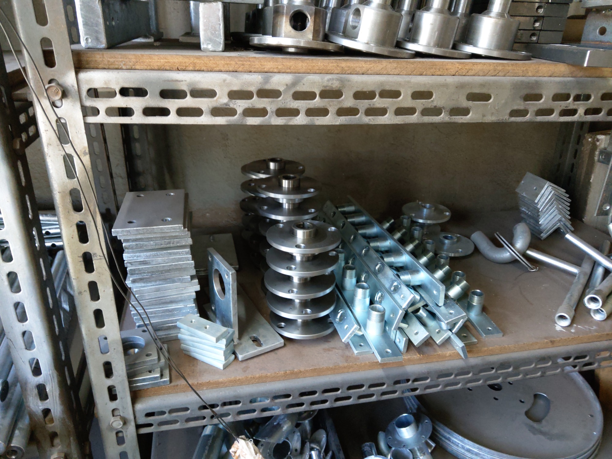 Metalowe elementy, w tym kołnierze, płyty i rury, ułożone na metalowych półkach regału warsztatowego.