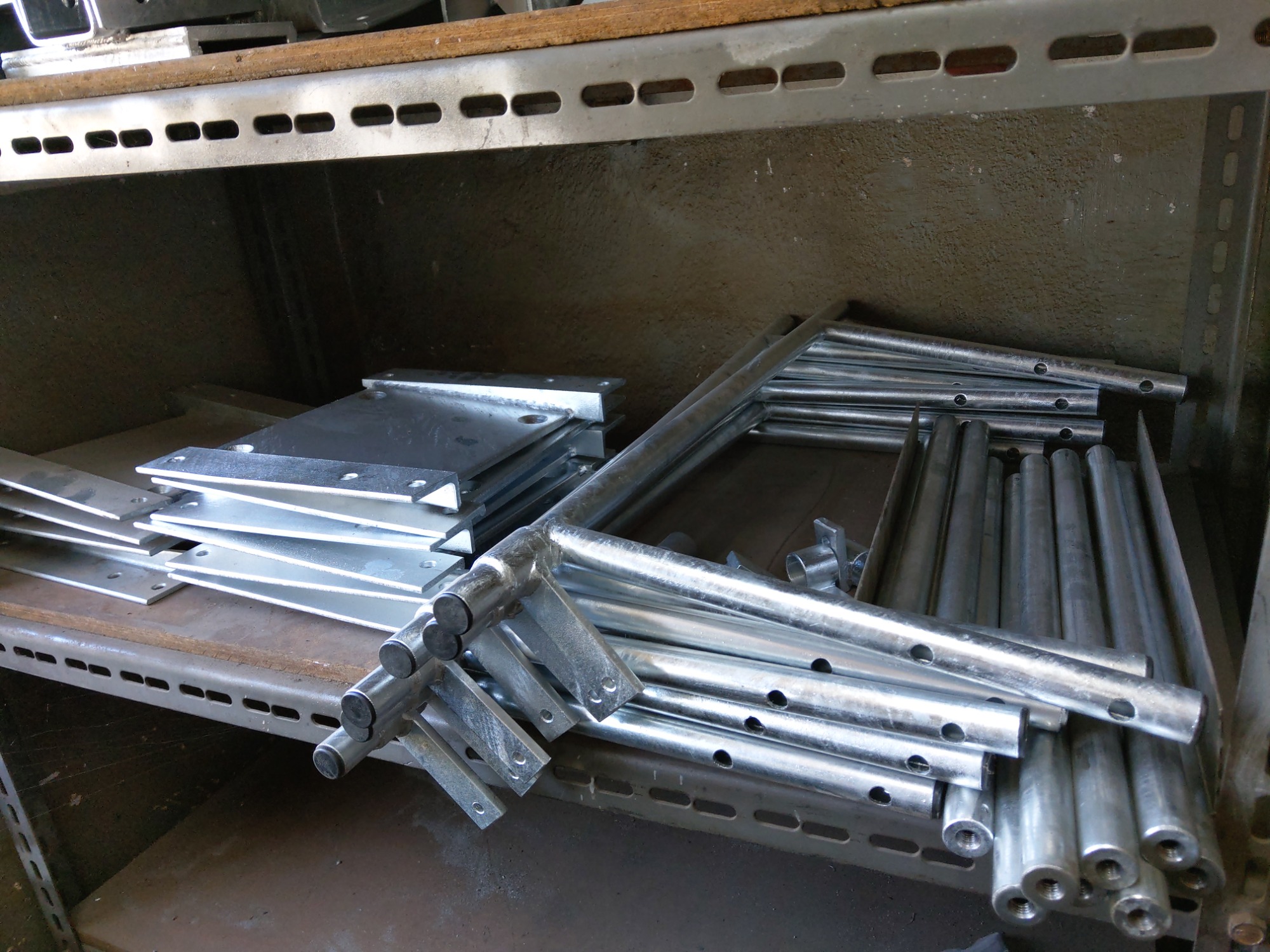 Stos ocynkowanych, metalowych elementów konstrukcyjnych z otworami i mocowaniami, ułożonych na półce w magazynie.