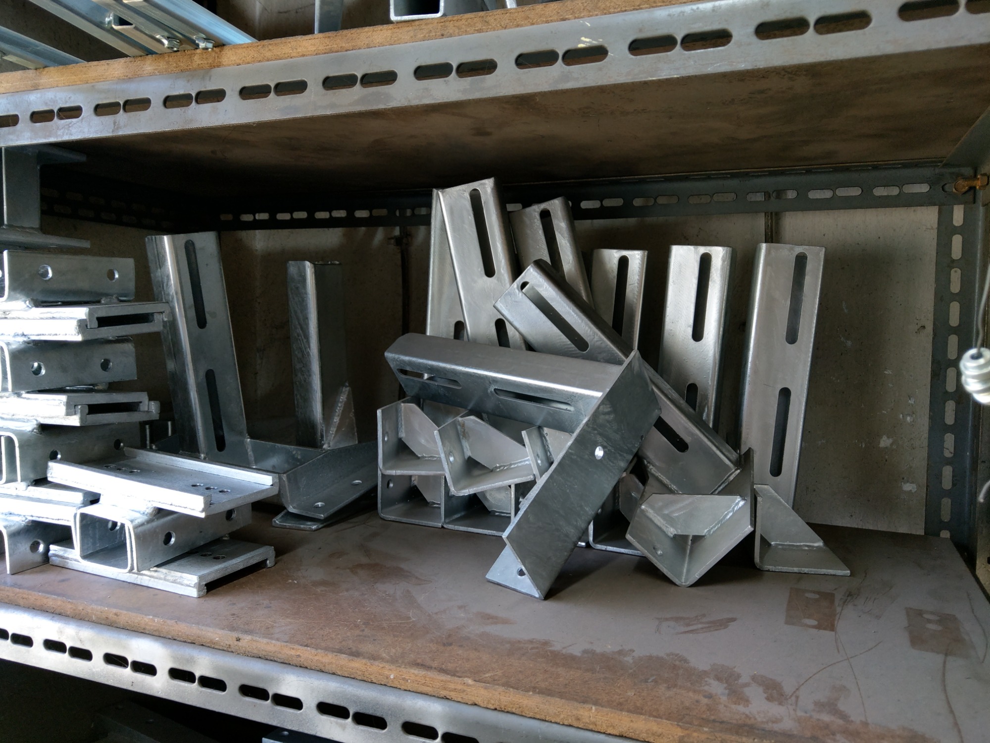 Metalowe wsporniki i profile o różnych kształtach, leżące na półce regału warsztatowego, niektóre z otworami i szczelinami, w surowym, srebrnym wykończeniu.