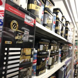 FHU Heda Adam Heda - Półka w sklepie budowlanym z ekspozycją palety kolorów fug marki Sopro oraz rzędami opakowań chemii budowlanej tej samej firmy, w odcieniach czerni i złota.