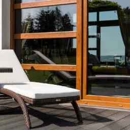 Leżanka z rattanu z białymi poduszkami na tarasie z desek kompozytowych, widok przez duże okno na ogród.