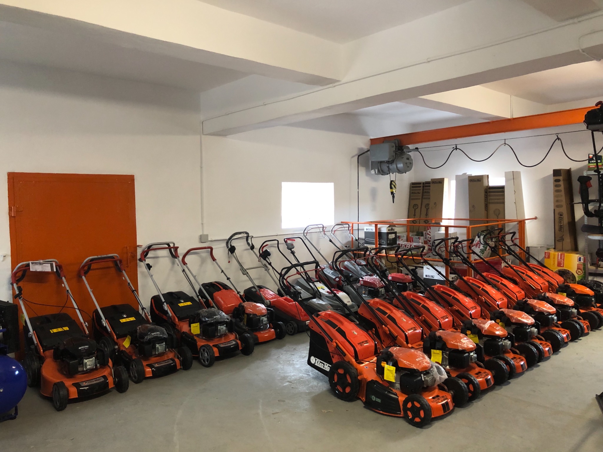 Rząd pomarańczowo-czarnych kosiarek do trawy ustawionych w magazynie, widoczne różne modele i detale konstrukcyjne, na podłodze szary beton, w tle białe ściany i pomarańczowe elementy konstrukcyjne.