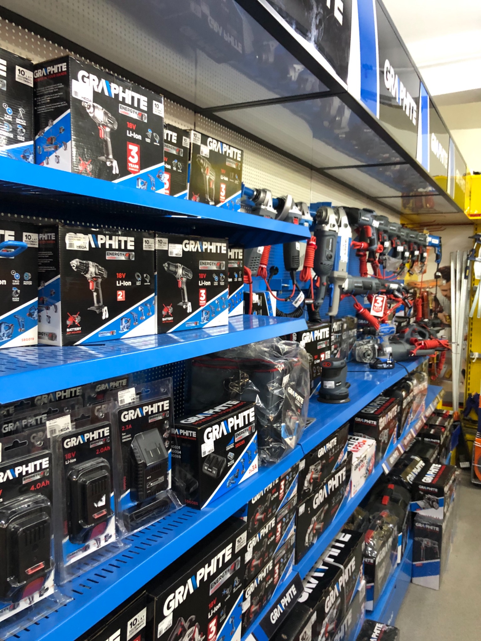 Półki w sklepie z narzędziami Graphite, prezentujące szeroki asortyment wiertarek akumulatorowych, szlifierek i akcesoriów, w tym baterie, wszystko w oryginalnych opakowaniach.