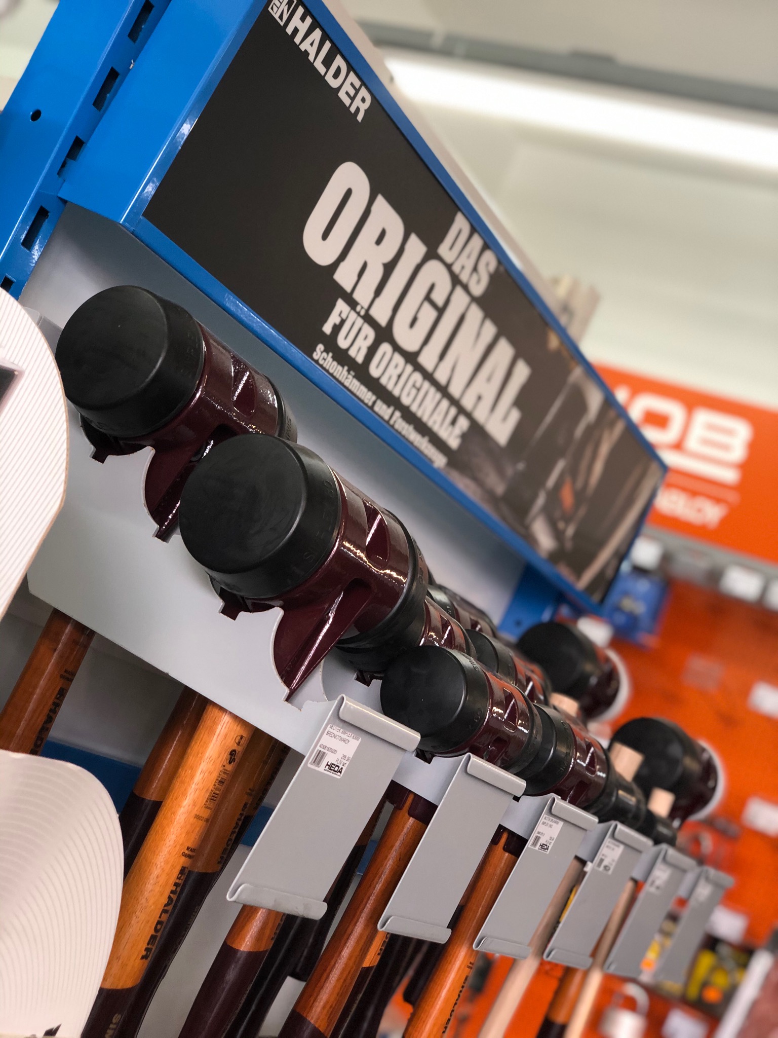 Ekspozycja młotków bezodrzutowych Halder na półce w sklepie, widoczne etykiety z cenami i logo producenta, tło w odcieniach pomarańczy.