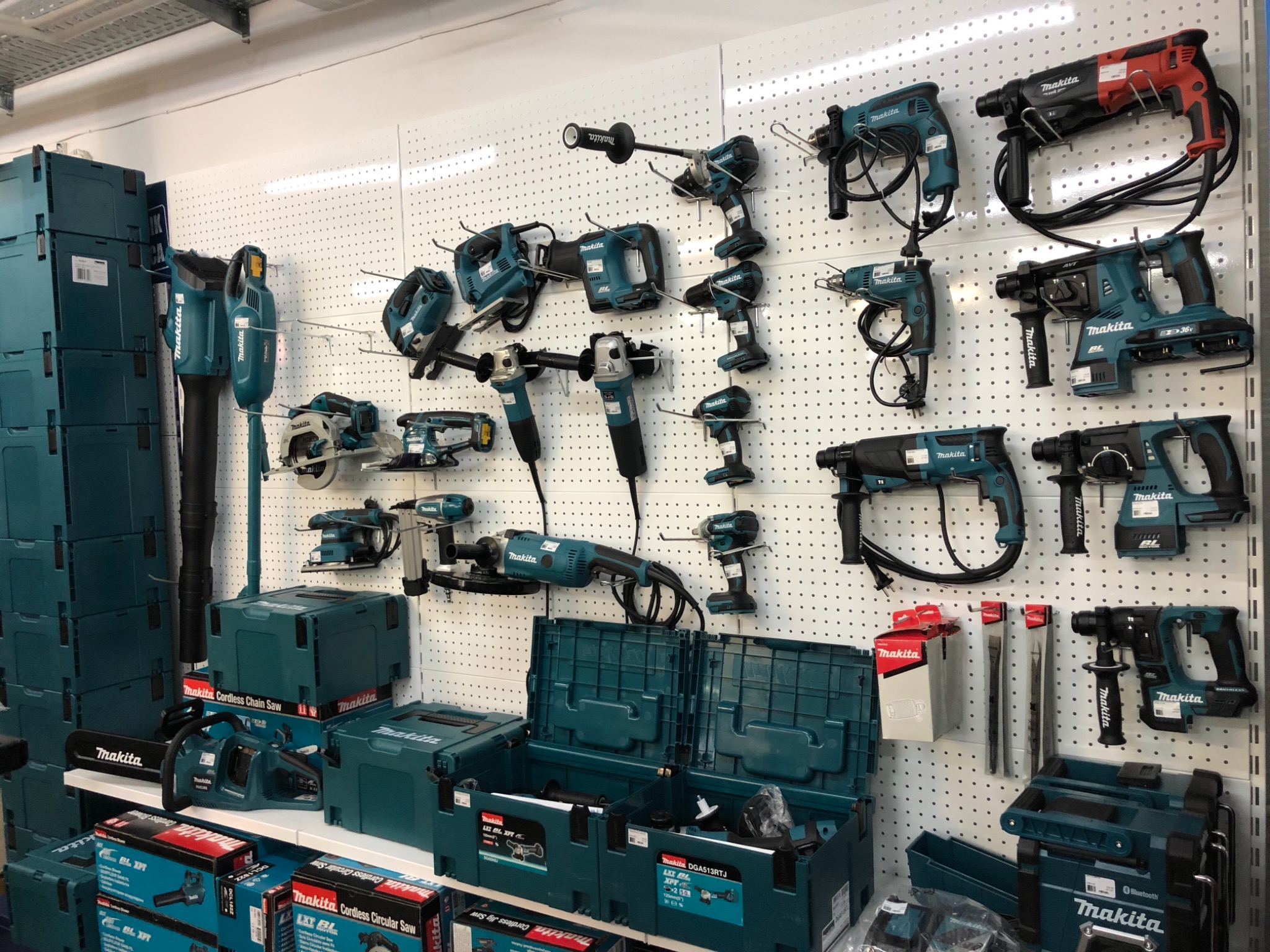 Ekspozycja narzędzi akumulatorowych marki Makita, w tym wiertarek, szlifierek, pilarek i akcesoriów, prezentowanych na perforowanej ścianie w sklepie, z licznymi opakowaniami i walizkami...