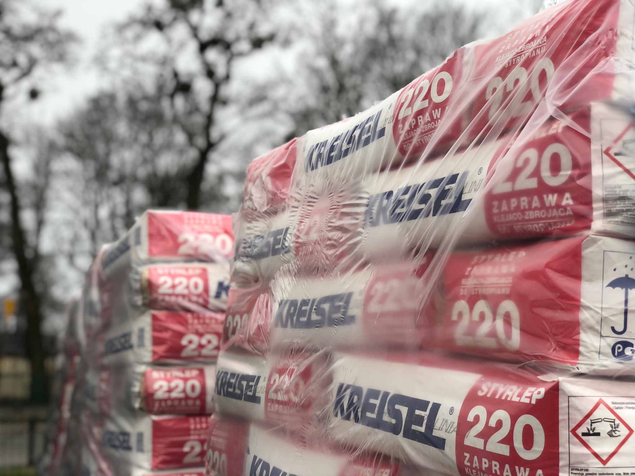Ułożone w stosy paczki zaprawy klejąco-zbrojącej do ociepleń, opakowane w folię, z widocznym logo producenta Kreisel i oznaczeniem Styrop 220, na tle rozmytych drzew.