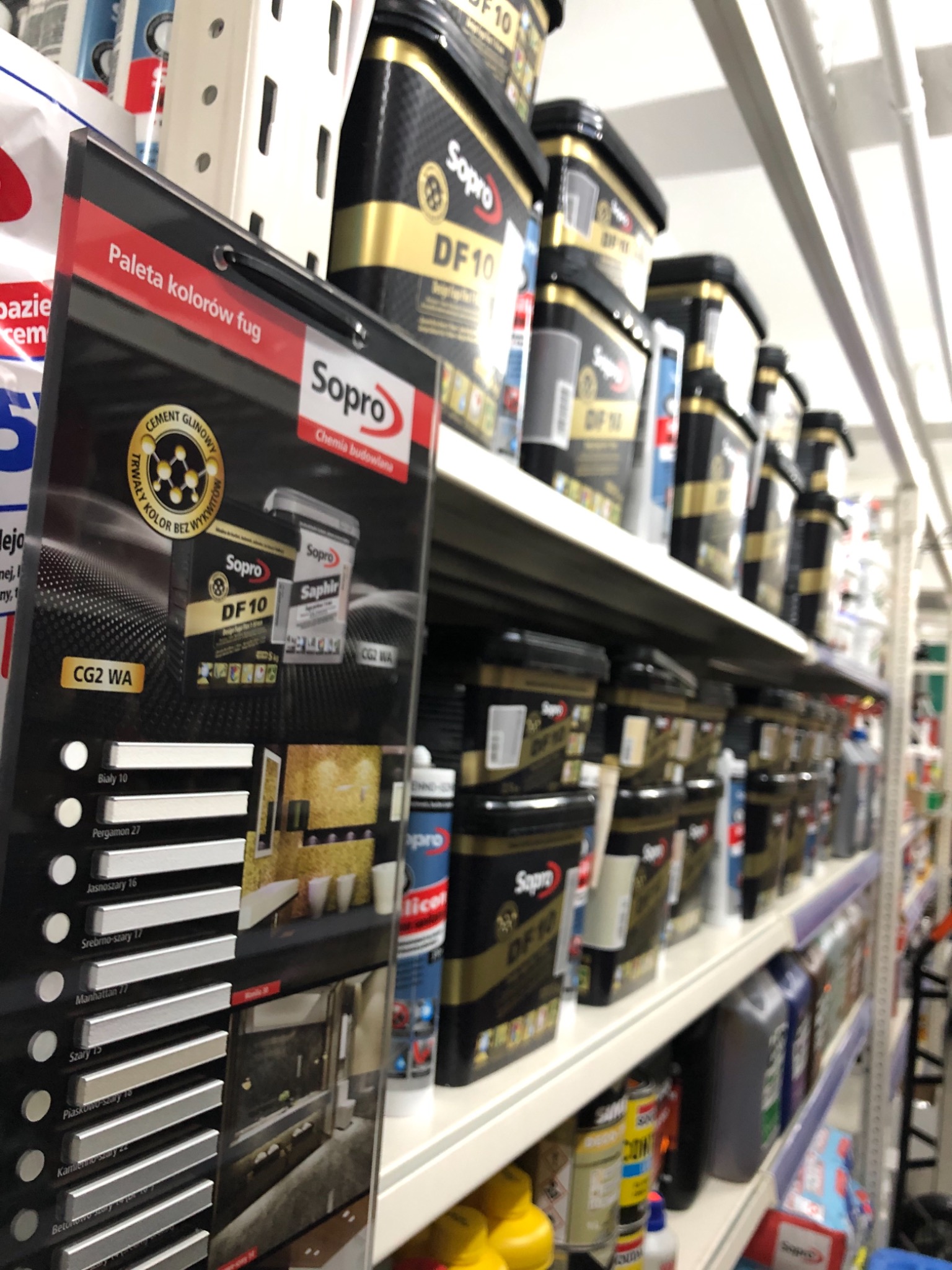 Półka w sklepie budowlanym z ekspozycją palety kolorów fug marki Sopro oraz rzędami opakowań chemii budowlanej tej samej firmy, w odcieniach czerni i złota.