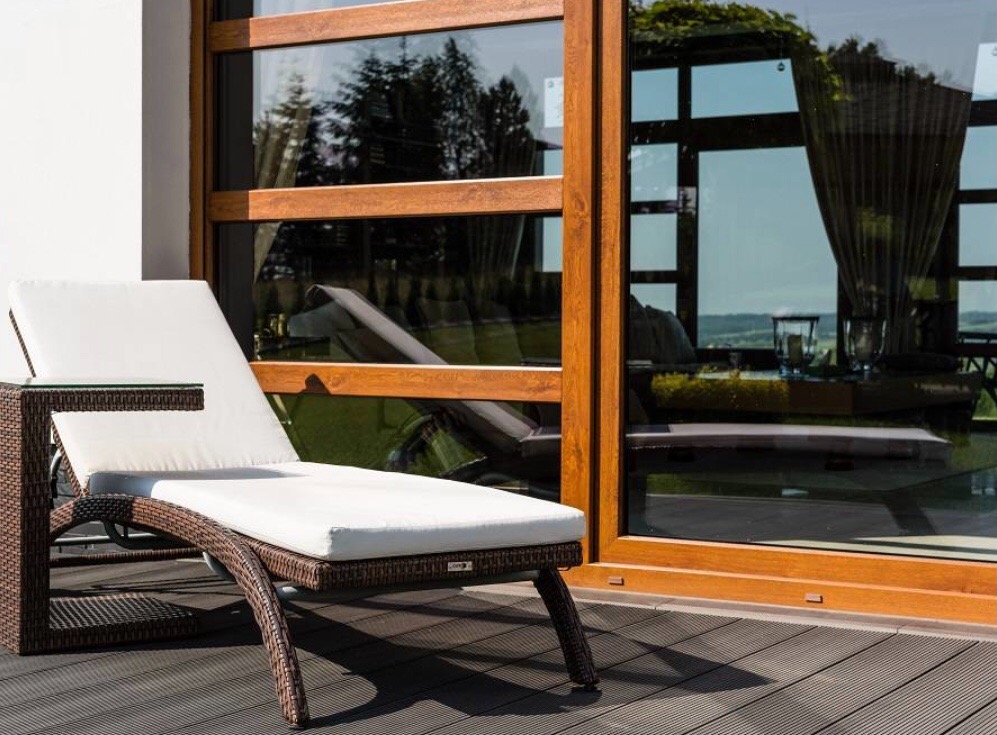Leżanka z rattanu z białymi poduszkami na tarasie z desek kompozytowych, widok przez duże okno na ogród.