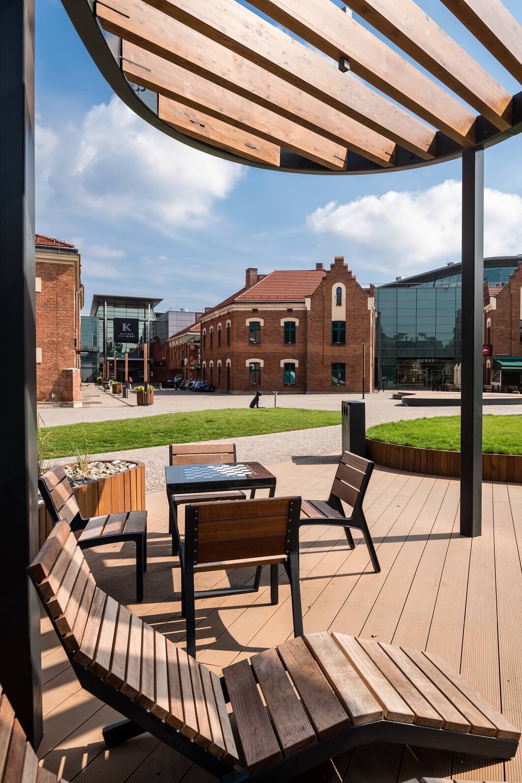 Zewnętrzny taras z drewnianą podłogą i meblami wypoczynkowymi, widok na budynki z czerwonej cegły i nowoczesną architekturę w tle, błękitne niebo z chmurami.