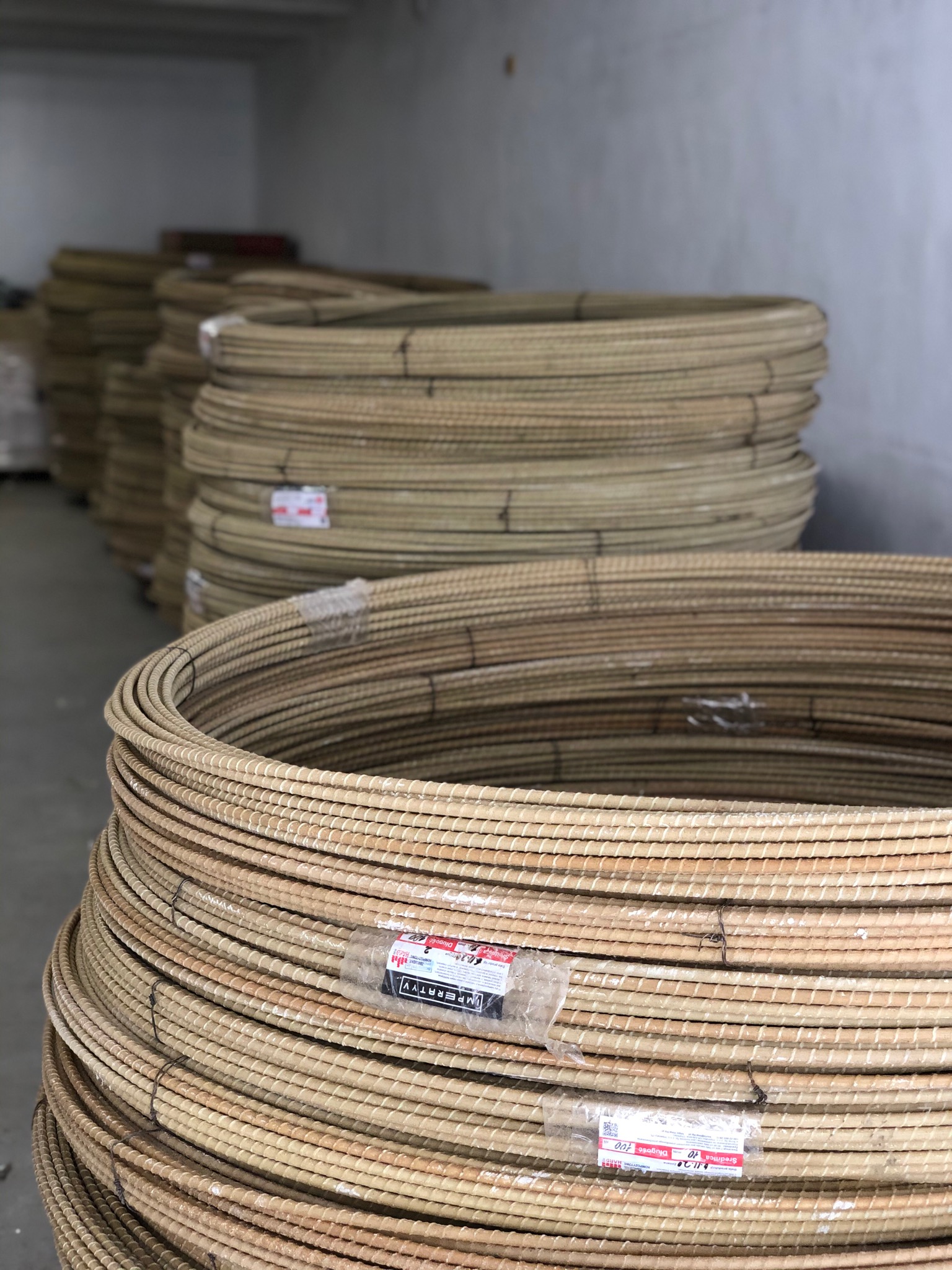 Zestaw zwojów kompozytowej stali zbrojeniowej w magazynie, widoczne etykiety producenta i szary beton na ścianach w tle.