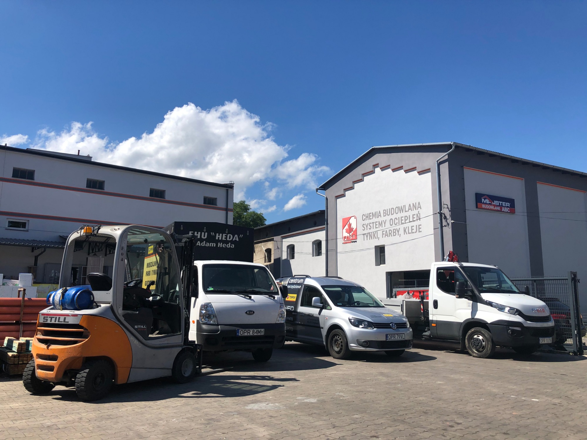 Plac firmy budowlanej z wózkiem widłowym Still, samochodami dostawczymi Kia i Iveco oraz budynkiem z reklamami chemii budowlanej i systemów ociepleń.