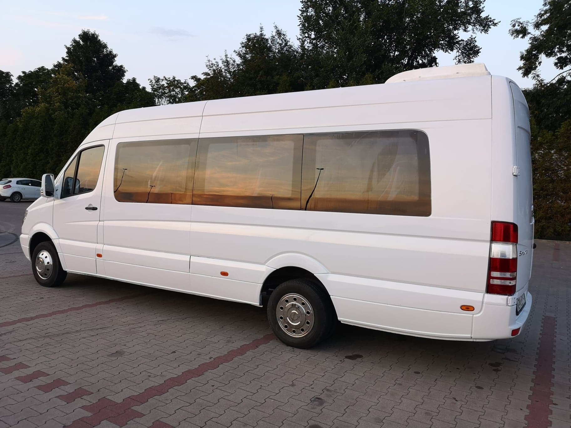 Biały, długi bus Mercedes-Benz Sprinter z dużymi oknami, zaparkowany na kostce brukowej, z wieczornym odbiciem nieba w oknach.