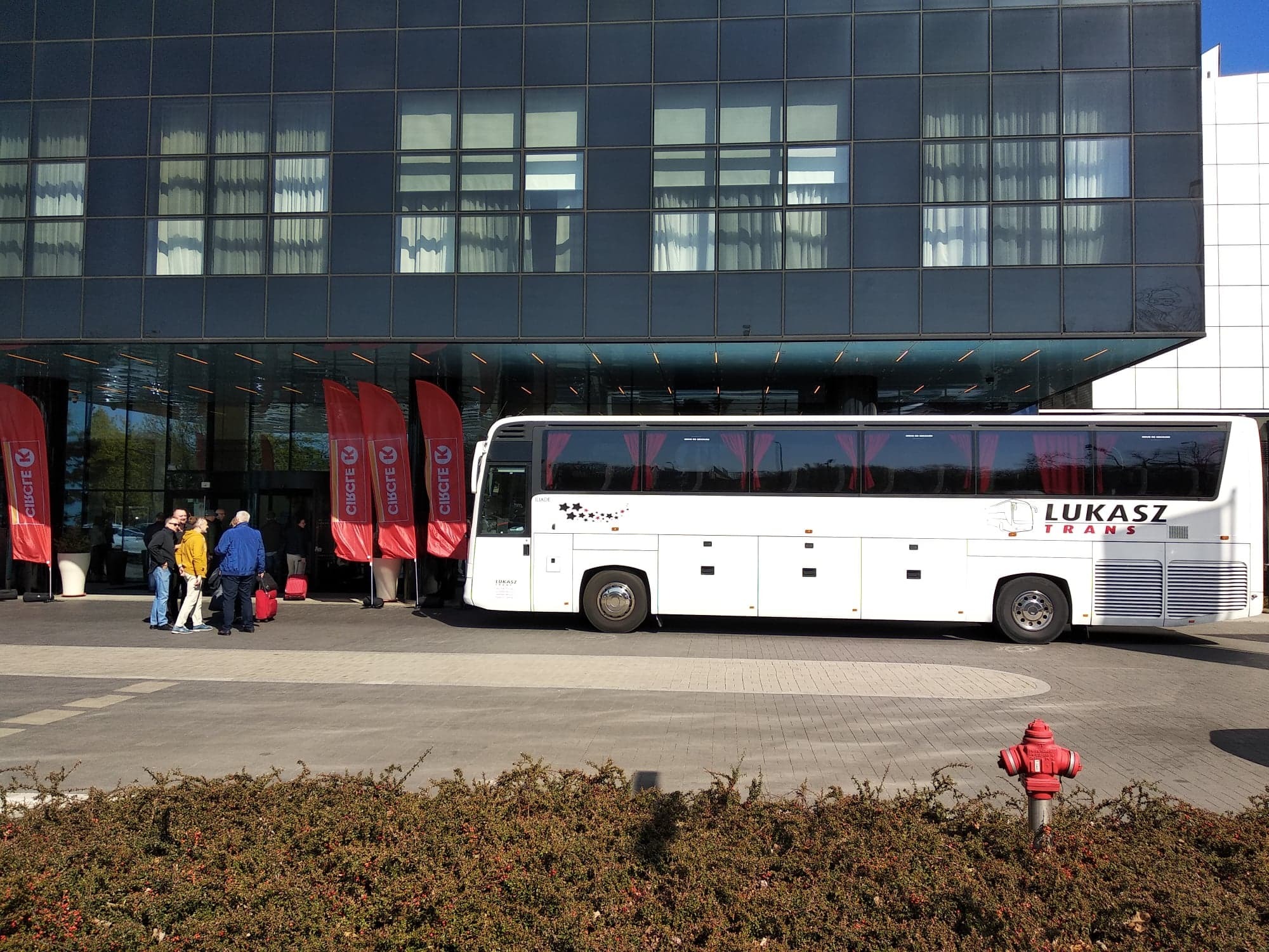Biały autokar firmy Łukasz Trans zaparkowany przed nowoczesnym budynkiem z ciemną szklaną fasadą, grupa osób z bagażem przy wejściu, czerwone flagi z logo, hydrant na pierwszym planie.