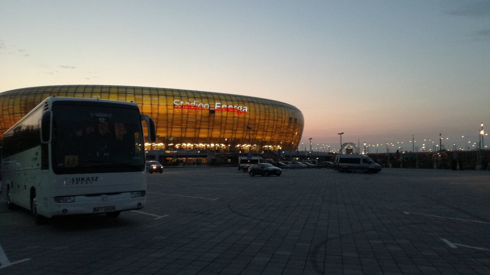 Biały autokar firmy Łukasz Trans przed Stadionem Energa w Gdańsku, wieczorne oświetlenie i zaparkowane auta.