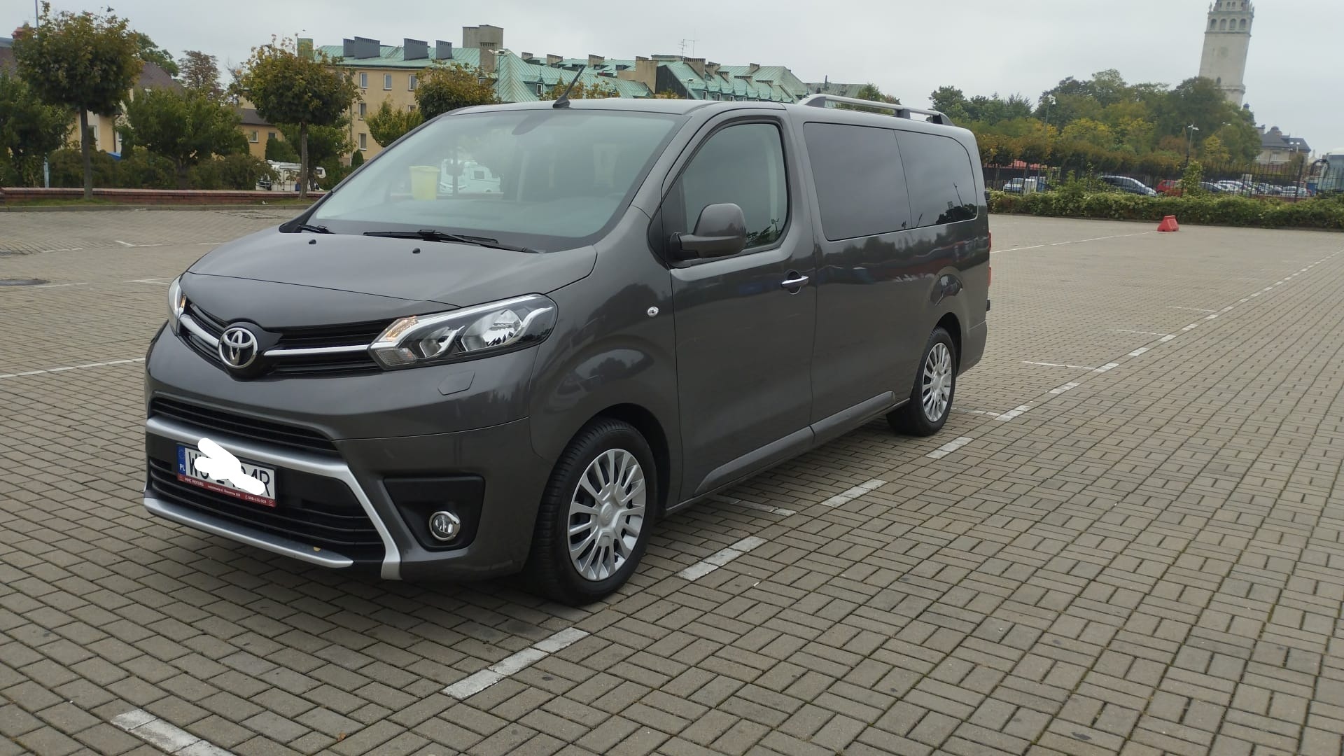 Szary van Toyota Proace Verso z relingami dachowymi, zaparkowany na parkingu z kostki brukowej, z wieżą w tle.