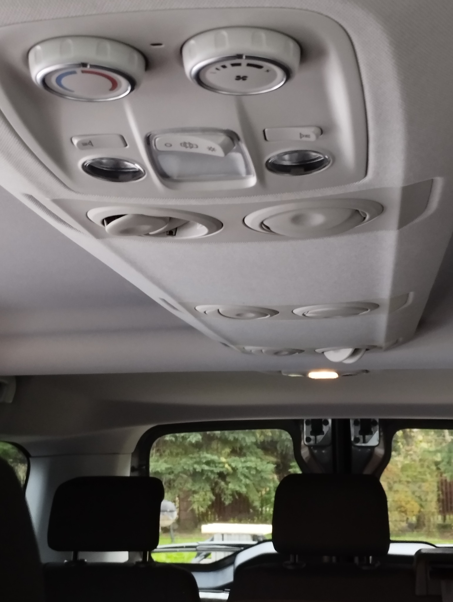 Panel sterowania klimatyzacją i nawiewami w przestrzeni pasażerskiej samochodu, widok z tyłu pojazdu, z widocznym fragmentem okna i drzewami na zewnątrz.