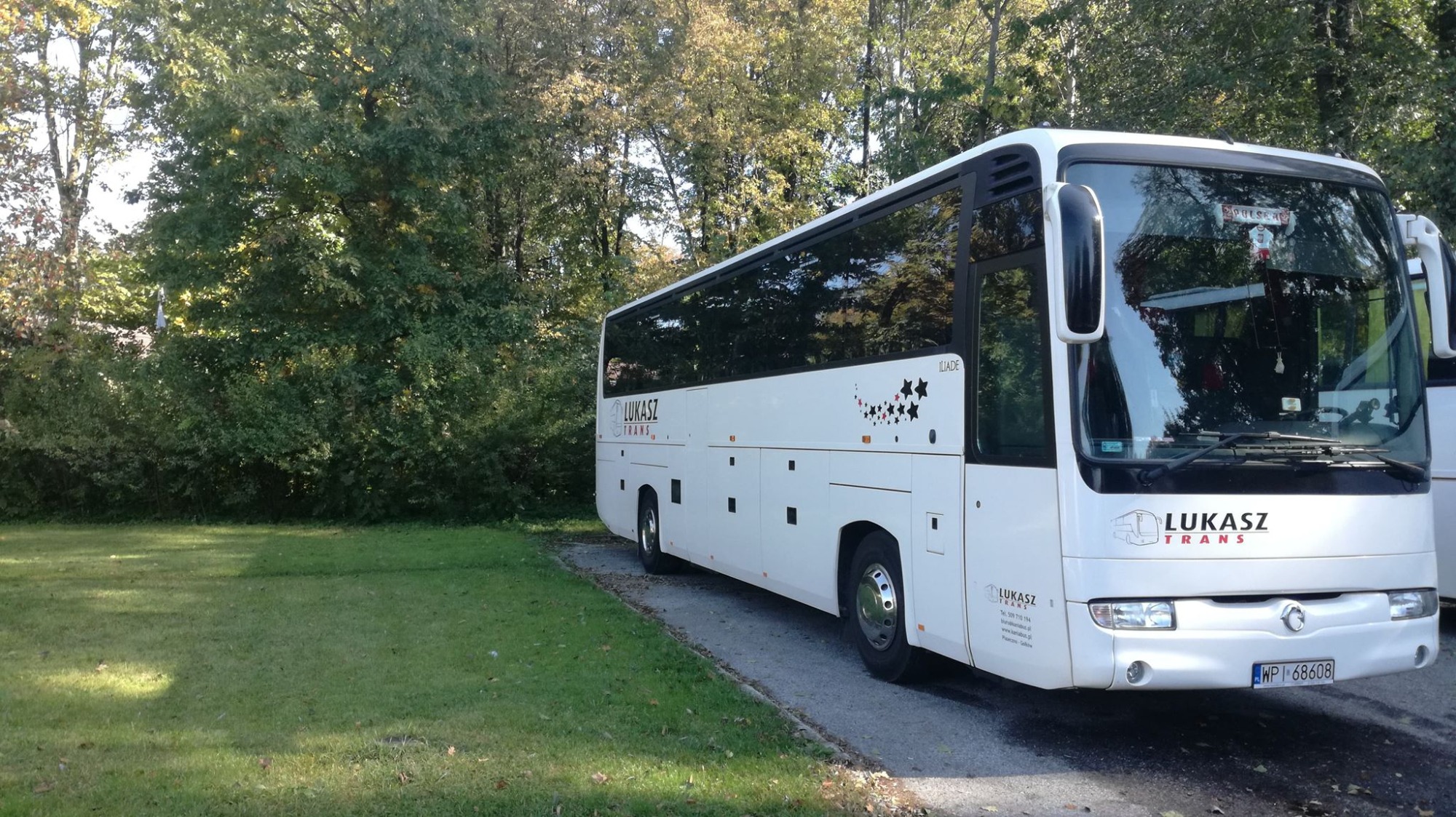 Biały autokar firmy Łukasz Trans zaparkowany na trawie obok drzew, z logo firmy na przedniej szybie i boku pojazdu, oraz dekoracyjnymi gwiazdkami na boku.