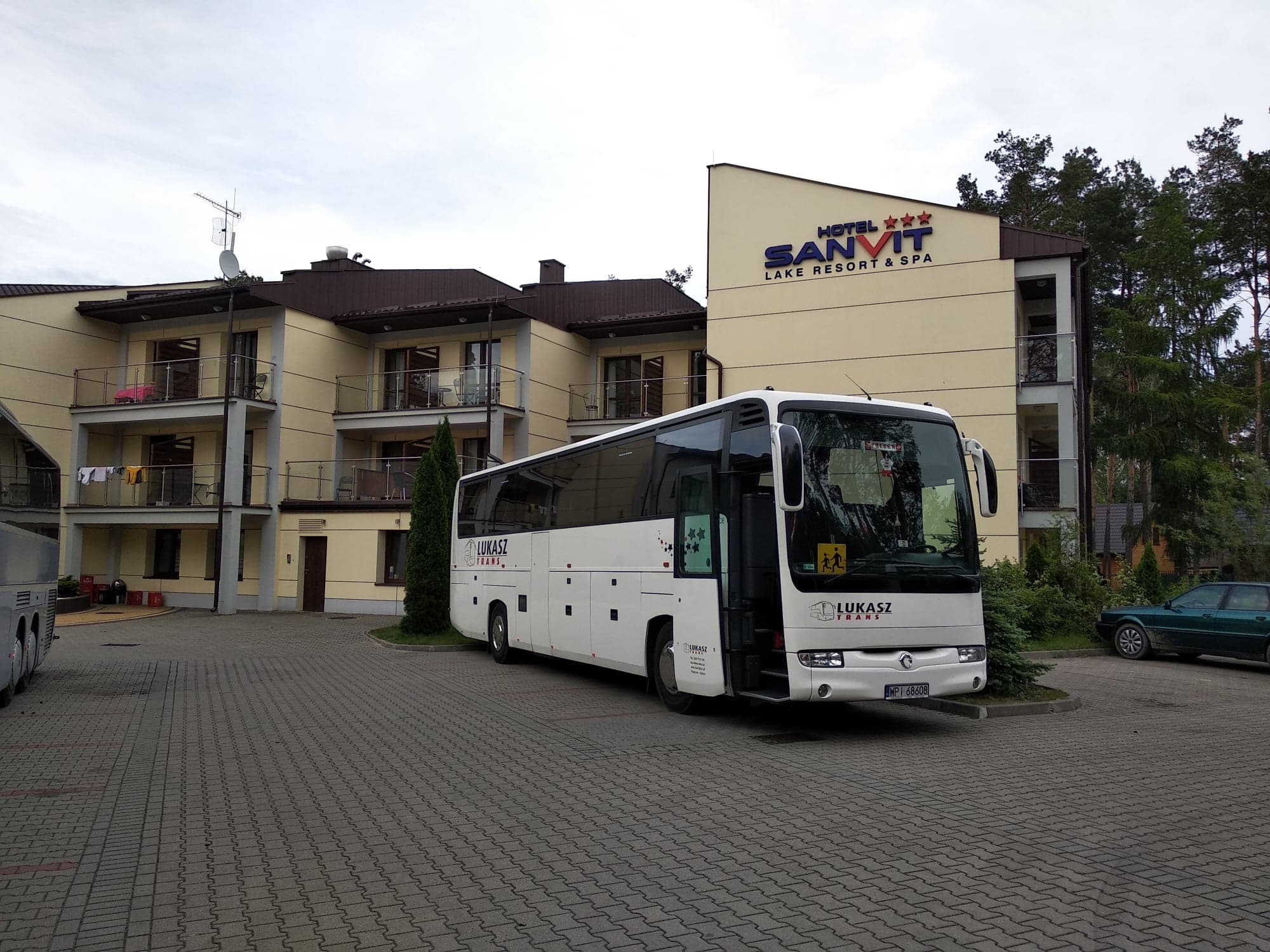 Autokar firmy Łukasz Trans zaparkowany przed hotelem Sanvit Lake Resort & Spa z widocznym znakiem ostrzegawczym przewozu dzieci.