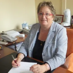 Kobieta w okularach i marynarce w paski siedzi za biurkiem, trzyma długopis i podpisuje dokument. Na biurku komputer, telefon i inne akcesoria biurowe.