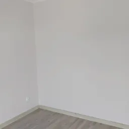 Jasnoszare panele podłogowe ułożone w rogu pokoju z jasnoróżowymi ścianami i białym sufitem, widoczny kontakt elektryczny.