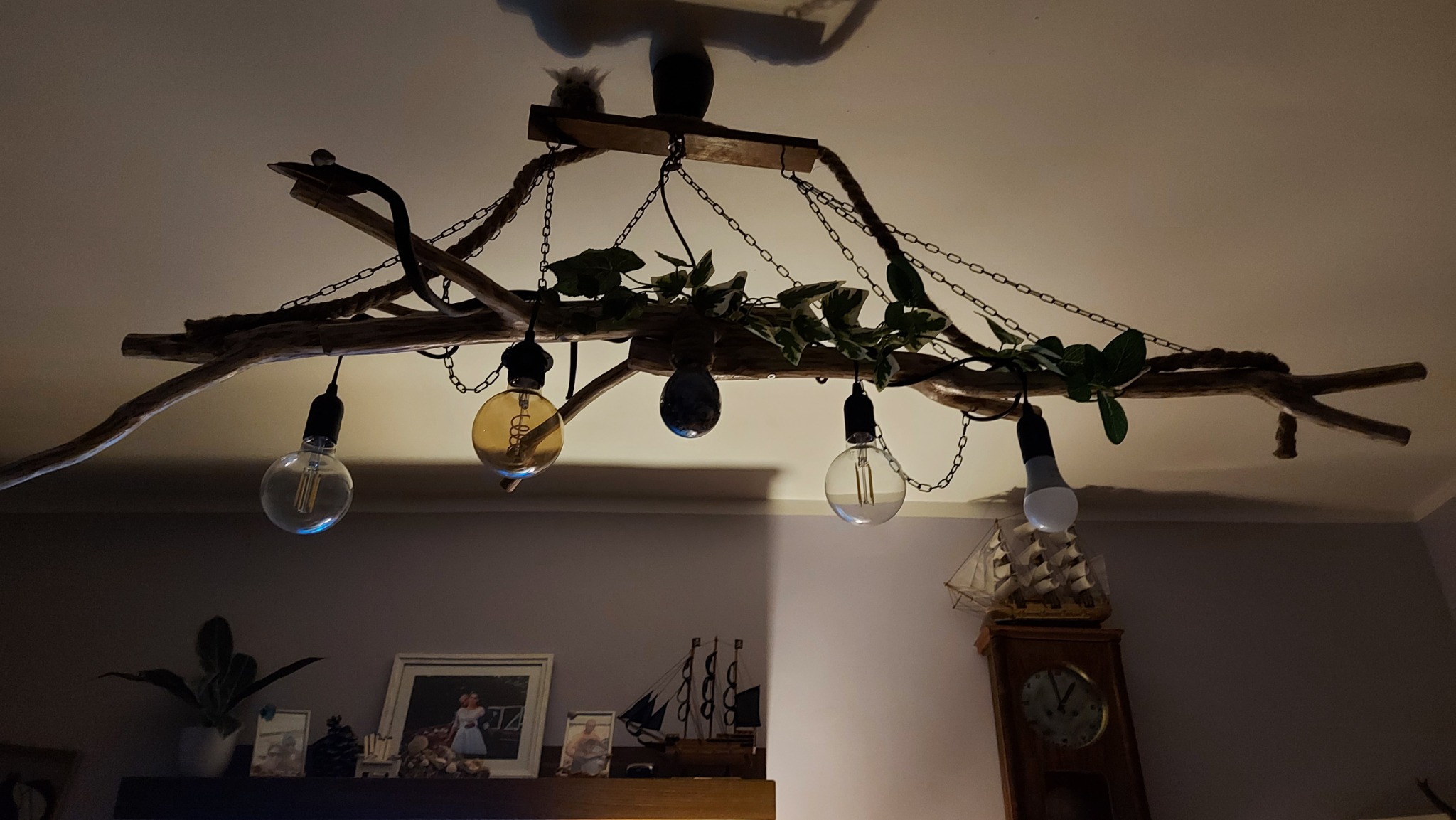 Dekoracyjna lampa sufitowa w stylu rustykalnym w Lubinie: gałąź drzewa z łańcuchami, żarówkami Edisona i sztucznymi liśćmi, oświetlająca półkę z dekoracjami.