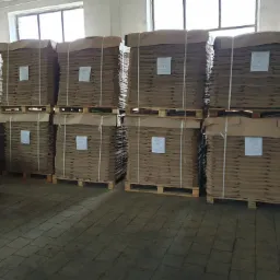 Stosy arkuszy tektury falistej, ułożone na drewnianych paletach, zabezpieczone taśmą i przykryte papierem, w hali magazynowej z betonową podłogą i oknami w tle.