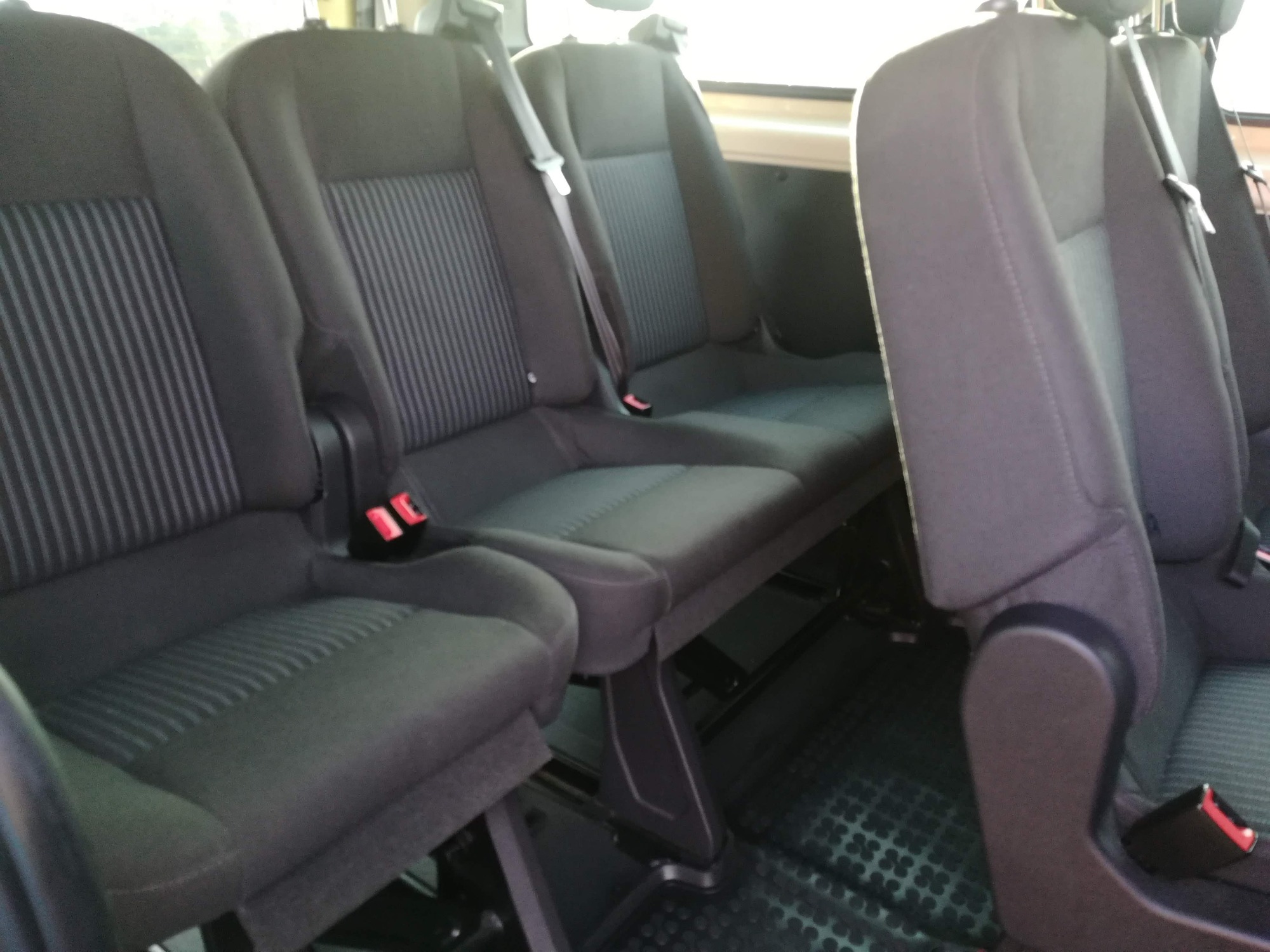 Wnętrze busa z trzema rzędami ciemnoszarych foteli z pasami bezpieczeństwa i wzorzystą tapicerką, widok z perspektywy pasażera.