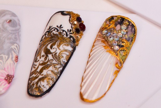 Precyzyjne zbliżenie na trzy sztuczne paznokcie z unikalnymi zdobieniami: czarno-złoty ornament na białym tle z czerwonymi i złotymi kryształkami, białe prążki z opalizującymi kryształkami...