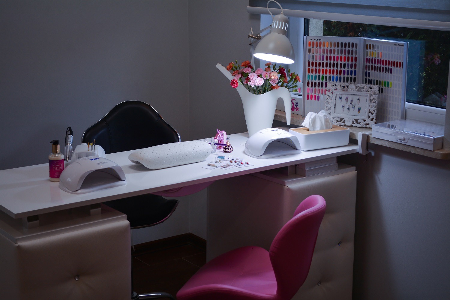 Stanowisko pracy w salonie kosmetycznym: białe biurko z lampą UV, poduszką pod dłonie, frezarką, lakierami, próbnikami kolorów i kwiatami w dzbanku.