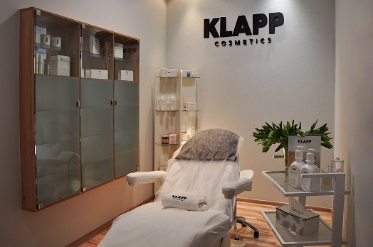 Jasne wnętrze gabinetu kosmetycznego z fotelem zabiegowym, szafkami ekspozycyjnymi z produktami Klapp Cosmetics i stolikiem z kosmetykami oraz zielonymi roślinami.