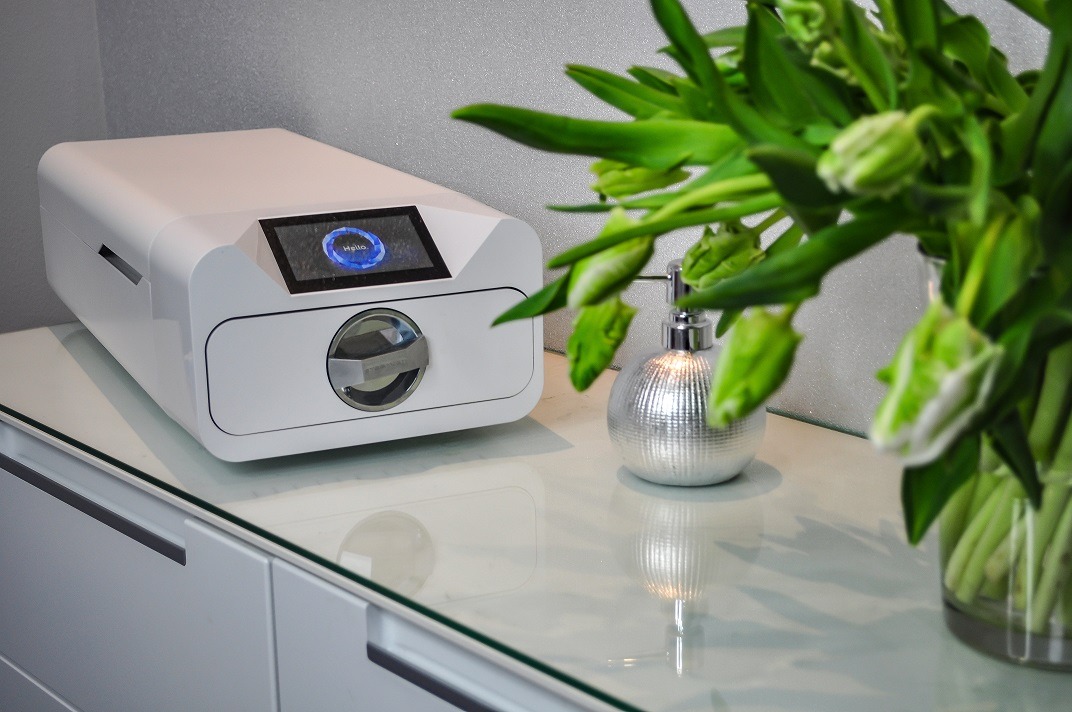 Nowoczesny, biały sterylizator kosmetyczny z wyświetlaczem i chromowanymi elementami, umieszczony na białej szafce z blatem z refleksami, obok srebrnego dozownika mydła i wazonu z zielonymi...