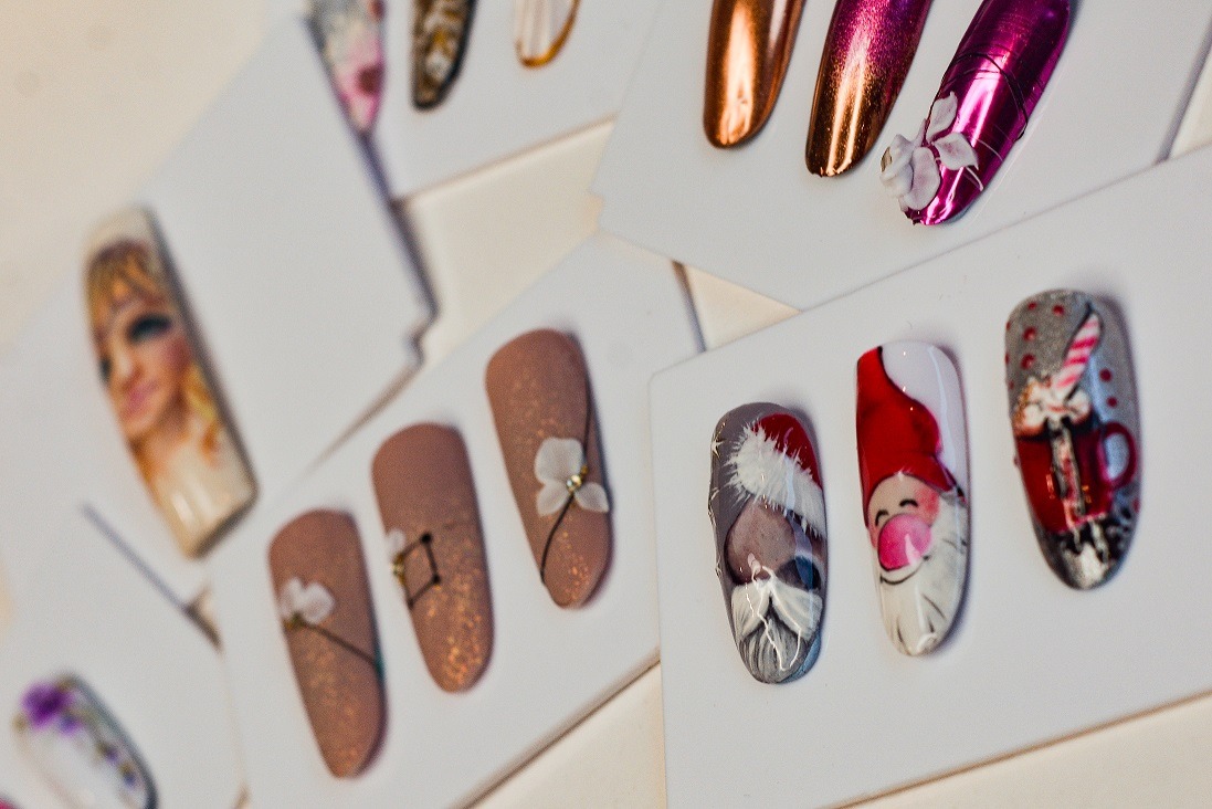 Prezentacja różnorodnych wzorów na sztucznych paznokciach, w tym motywy świąteczne Mikołaja i ozdoby kwiatowe, umieszczonych na białym tle.