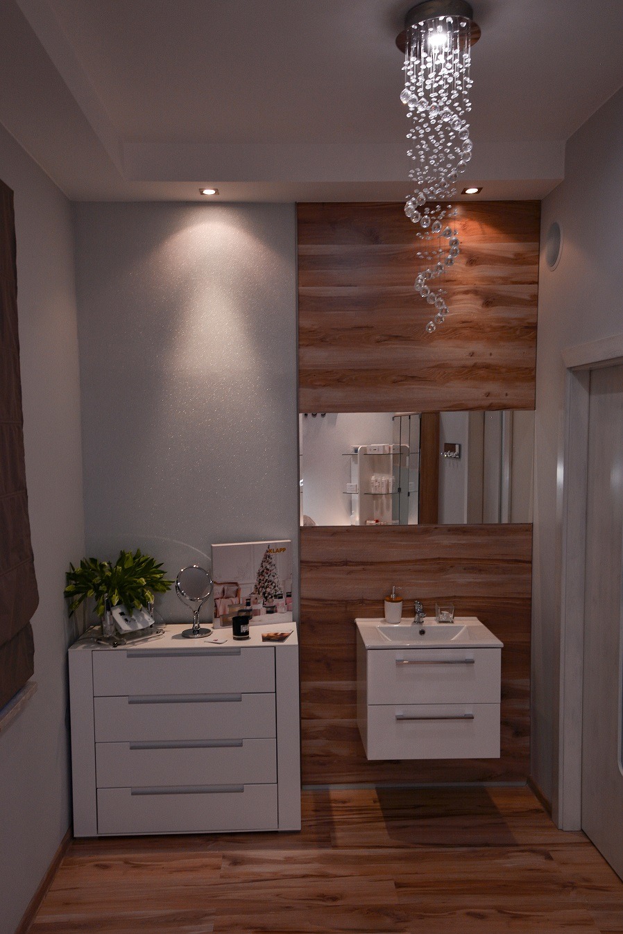 Nowoczesne wnętrze salonu kosmetycznego z białą komodą, umywalką i lustrem w drewnianej ramie, oświetlone dekoracyjnym kryształowym żyrandolem.