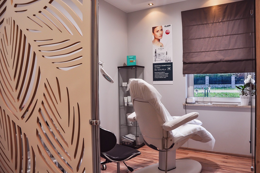 Jasne wnętrze gabinetu kosmetycznego z fotelem zabiegowym przykrytym pokrowcem, dekoracyjną ścianką z motywem liści i plakatem reklamowym na ścianie.