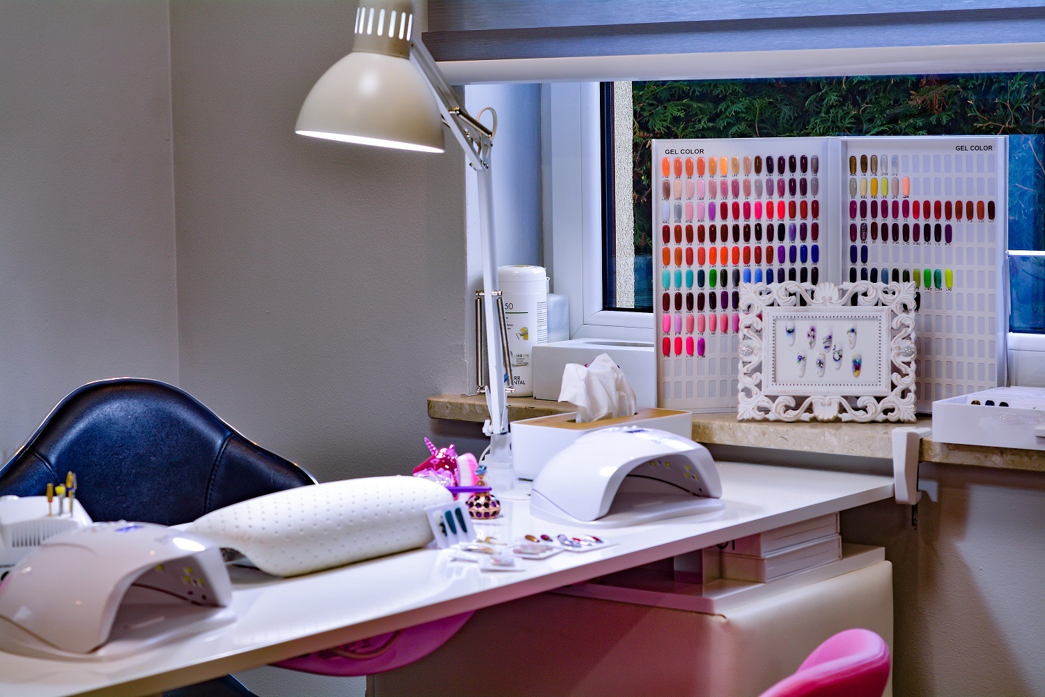 Stanowisko do stylizacji paznokci z lampami UV, poduszką, paletą kolorów żeli i ramką ze wzorami paznokci, oświetlone lampką biurkową, z widokiem na zieleń za oknem.