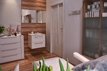 Jasne wnętrze gabinetu z drewnianymi akcentami, widoczne białe meble, umywalka z lustrem, przeszklona szafka z kosmetykami oraz fragment fotela zabiegowego z zielonymi roślinami w tle.