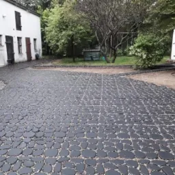 Czarna kostka brukowa ułożona w kształcie nieregularnych kamieni, oddzielona od trawnika pasem żwiru, na tle białego budynku gospodarczego z drewnianymi wrotami.