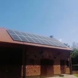 Panele słoneczne zamontowane na dachu ceglanego budynku gospodarczego z czerwoną dachówką, widoczne w słoneczny dzień.