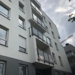 Nowoczesny budynek mieszkalny z balkonami zabezpieczonymi szklanymi balustradami z czarnymi ramami, widok z dołu na tle pochmurnego nieba, z żurawiem budowlanym w tle.