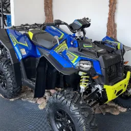 Niebiesko-żółty quad Polaris Scrambler XP 1000 na ekspozycji w salonie, otoczony dekoracyjnymi elementami imitującymi korę drzew.
