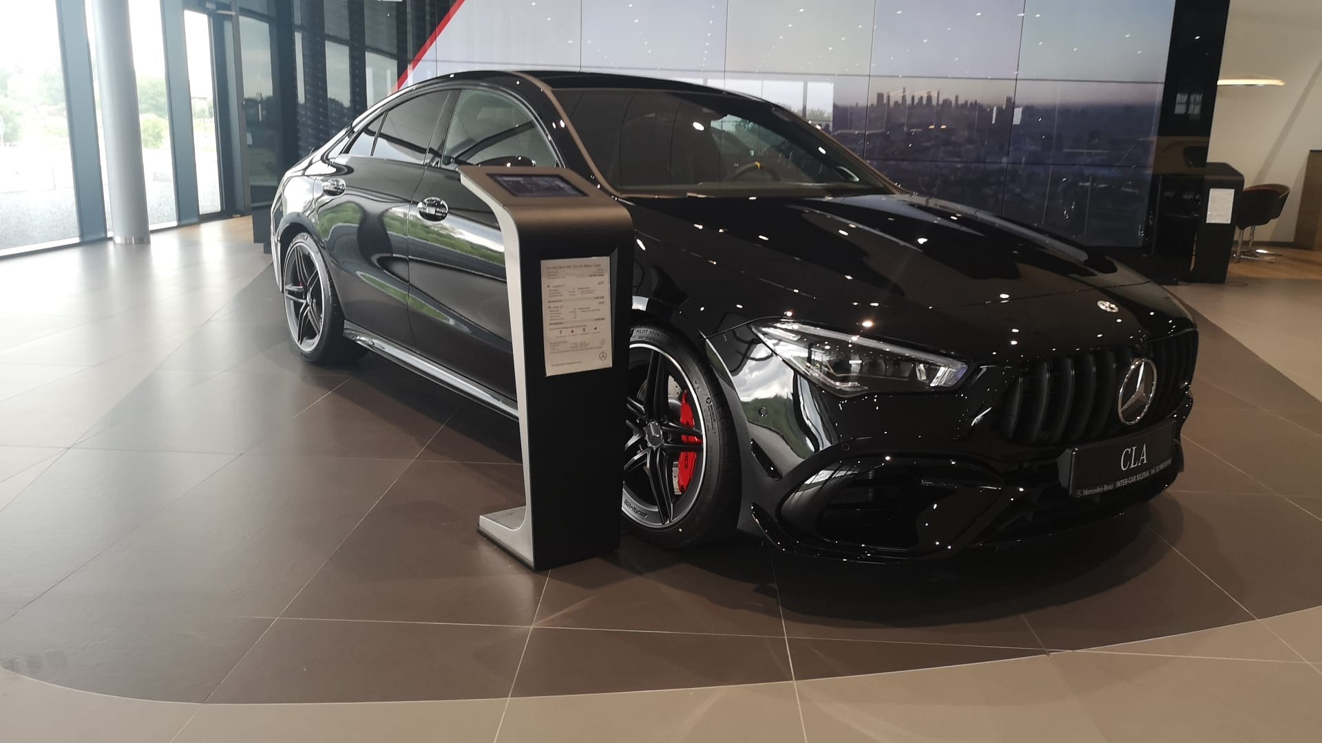 Czarny Mercedes-Benz CLA AMG z czerwonymi zaciskami hamulcowymi prezentowany w salonie samochodowym, obok interaktywny kiosk z informacjami o modelu.