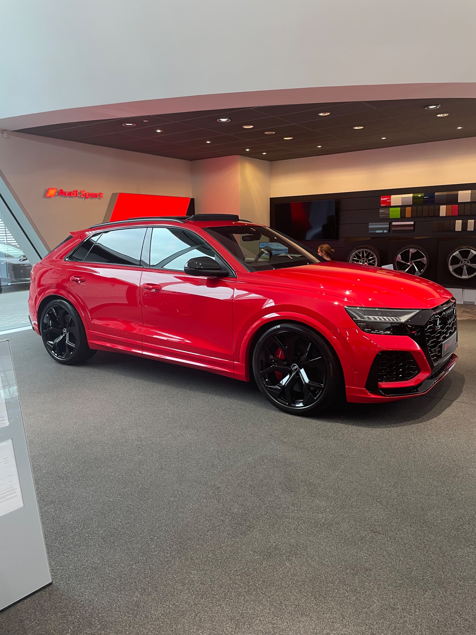 Czerwony samochód Audi RS Q8 z czarnymi felgami prezentowany w salonie samochodowym z logo Audi Sport w tle.