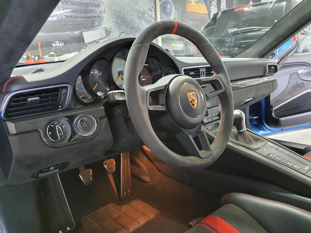 Wnętrze sportowego samochodu Porsche z szarą kierownicą obszytą czerwoną nicią, widoczne detale deski rozdzielczej z włókna węglowego i aluminiowe pedały.