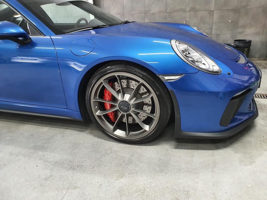 Błękitny sportowy samochód marki Porsche z widocznymi czerwonymi zaciskami hamulcowymi za szarymi felgami, uchwycony z niskiej perspektywy w czystym garażu.